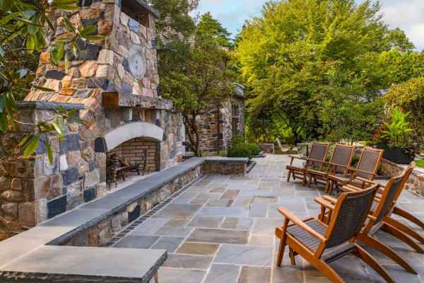 Outdoor living in Delaware County, PA features a patio with a stone fireplace and chimney, surrounded by elegant stone walls. Wooden lounge chairs with cushions face the hearth, while lush greenery creates a serene atmosphere. Perfect for enjoying nature's tranquility.