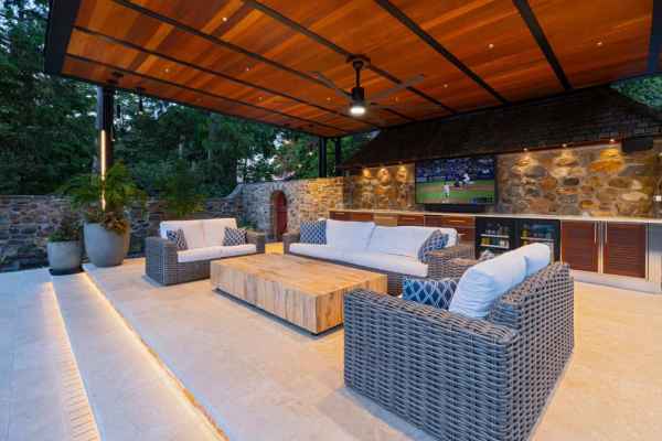 Outdoor patio featuring wicker sofas around a wooden table, with a stone wall backdrop and a TV showcasing the game. Ceiling fans whirl above, and lush greenery thrives. Stairs lead to this cozy outdoor living space in Delaware County, PA, under a pergola adorned with potted plants.