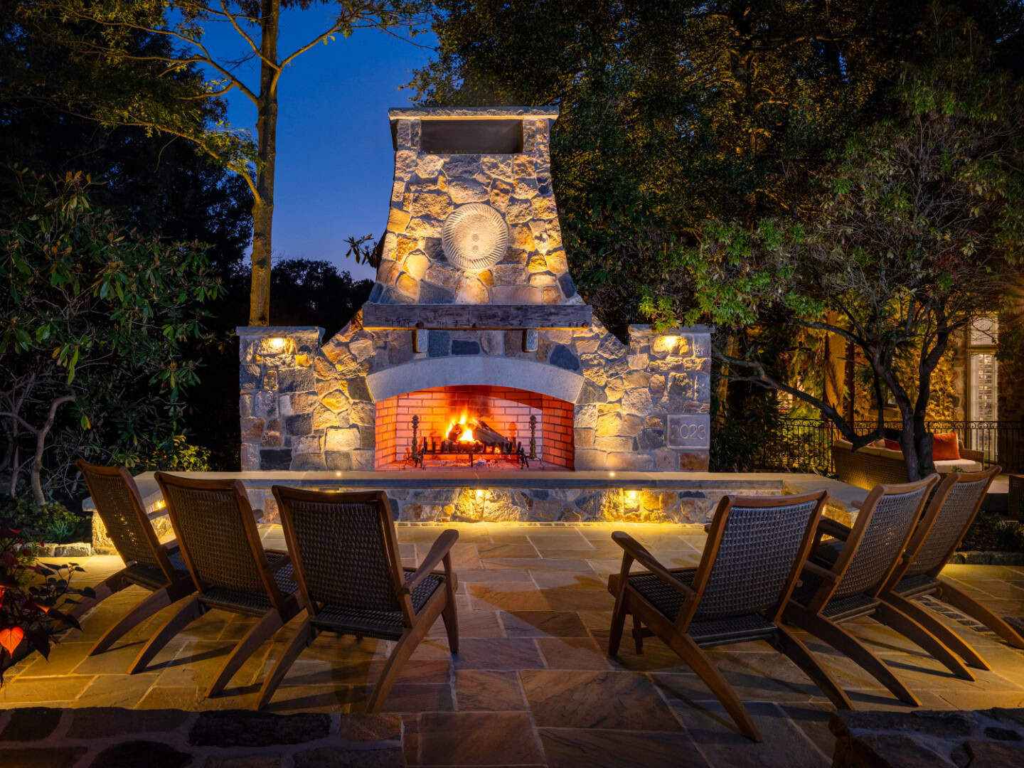 A cozy stone outdoor fireplace illuminates the night, casting a warm glow. Five wooden lounge chairs form a semicircle on the stone patio, embraced by lush greenery and the clear evening sky—a perfect example of outdoor living in Delaware County, PA.
