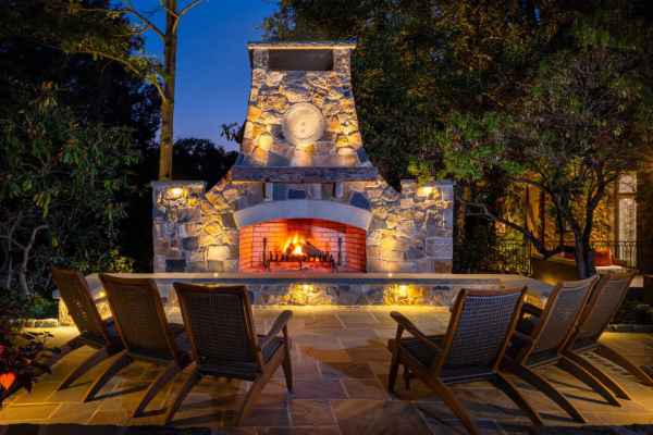 A cozy stone outdoor fireplace illuminates the night, casting a warm glow. Five wooden lounge chairs form a semicircle on the stone patio, embraced by lush greenery and the clear evening sky—a perfect example of outdoor living in Delaware County, PA.