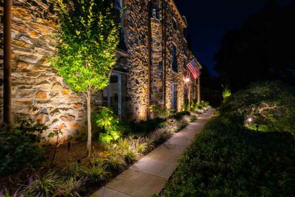 Stone house exterior at night, with warm lighting accentuating textured walls. A well-lit pathway with plants and artful hardscaping leads alongside the building. An American flag is displayed near the entrance, while expertly manicured bushes line the way—showcasing top-notch landscaping services in Delaware County PA.