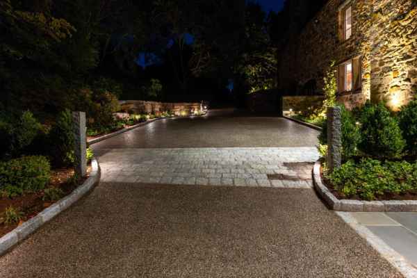 The illuminated driveway at night leads to a stone house, its warmly lit windows inviting you in. Expert landscaping services in Delaware County, PA have lined the path with green shrubs and accented it with stone columns against a backdrop of silhouetted trees.