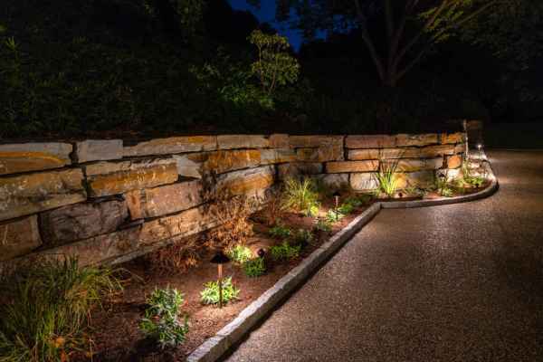 A curved stone wall with a garden bed is beautifully lit by small landscape lights at night. The paved path beside it invites exploration, while trees stand silhouetted against the dark sky, showcasing expert hardscaping services in Delaware County, PA.