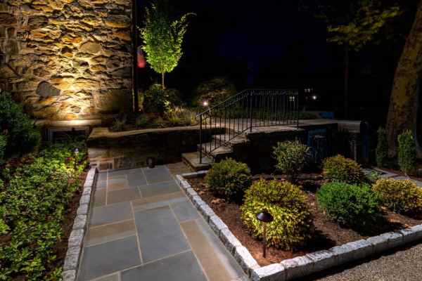 A nighttime garden scene with a stone walkway surrounded by neatly trimmed bushes and shrubs showcases the beauty of outdoor living in Delaware County, PA. A small staircase with a metal railing leads to a raised area. Ambient lighting highlights the stone wall and a small tree.