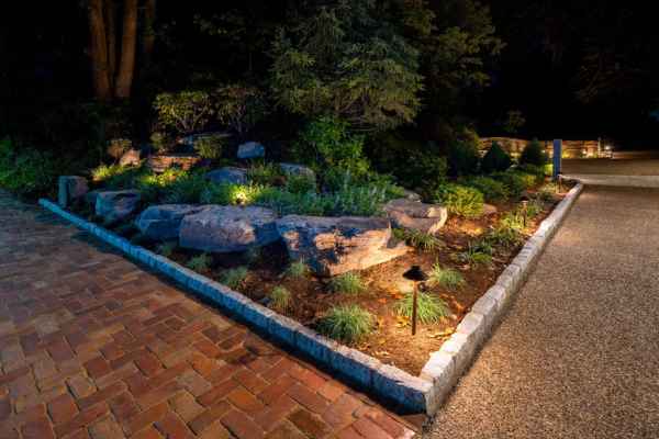 A landscaped garden at night with spotlights illuminating rocks and plants is bordered by a stone edge, adjacent to a brick pathway. Trees and shrubs provide a dark contrast in the background, showcasing the beauty of outdoor living in Delaware County, PA.