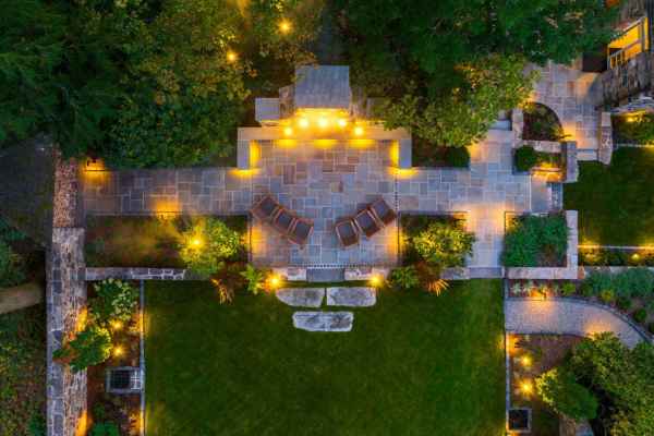 Aerial view of a beautifully lit patio and garden at night. The stone patio features wooden chairs and glowing lights outlining the area. Expert hardscaping services in Delaware County, PA have enhanced the lush green grass and pathways, with trees and plants providing a natural border.