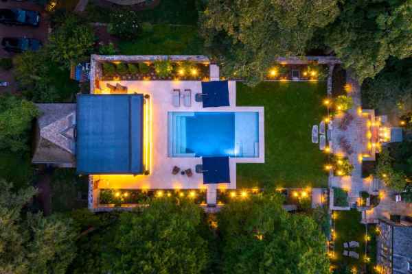 Aerial view of a lit-up backyard at night featuring a swimming pool surrounded by patio furniture and umbrellas. The space, expertly crafted with landscaping services in Delaware County, PA, is bordered by trees and a lawn with decorative lights, adjacent to a building and pathways.