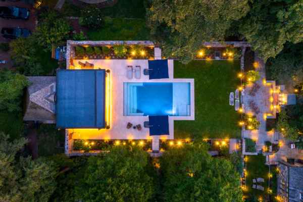 Aerial view of a lit-up backyard with a rectangular swimming pool, surrounded by a patio and lush greenery. Lounge chairs and umbrellas line the pool. String lights illuminate the garden, showcasing outdoor living Delaware County PA, and creating a cozy, inviting atmosphere.