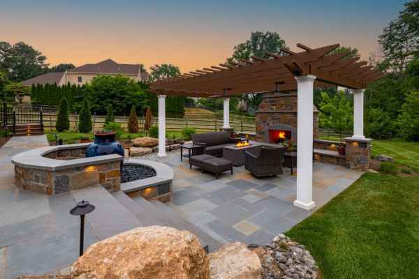 Luxurious outdoor patio at sunset with a stone fire pit and comfortable wicker chairs under a large pergola. The lush setting is enhanced by expert hardscaping services in Delaware County, PA, boasting beautifully arranged rocks and hedges with a spacious lawn beyond.