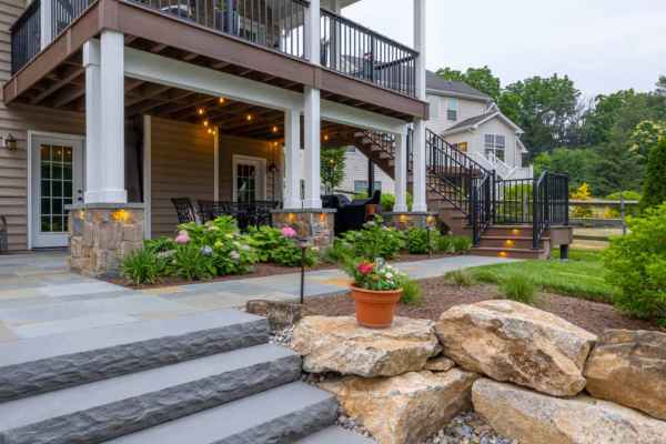 A two-story patio with a dining area beneath wooden decking showcases the best of outdoor living Delaware County PA. Steps, flanked by landscaped gardens with rocks and potted plants, lead up to the deck. Embedded lighting in the stairs illuminates this inviting space, with a house in the background.
