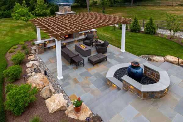 Aerial view of a landscaped backyard with a pergola-covered patio, featuring outdoor seating, a fireplace, and a fountain. Designed for optimal outdoor living in Delaware County, PA, the patio is surrounded by lush grass, trees, and flower beds with rocks.