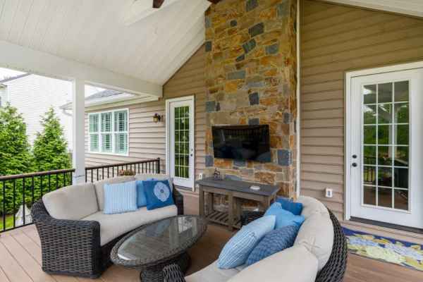 Outdoor living in Delaware County, PA: A cozy patio with wicker furniture, blue cushions, and a glass-top coffee table. A stone fireplace with a mounted TV adorns the light brown siding. French doors lead inside, while a ceiling fan and railing enhance the setting. Ideal for enjoying hardscaping services nearby.