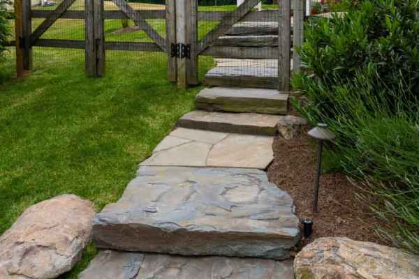 Stone steps lead up a grassy slope to a wooden garden gate, an inviting entryway designed with expert landscaping services in Delaware County, PA. A white pergola graces the background, while the path is lined with large rocks and lush green plants, enhancing your outdoor living experience.
