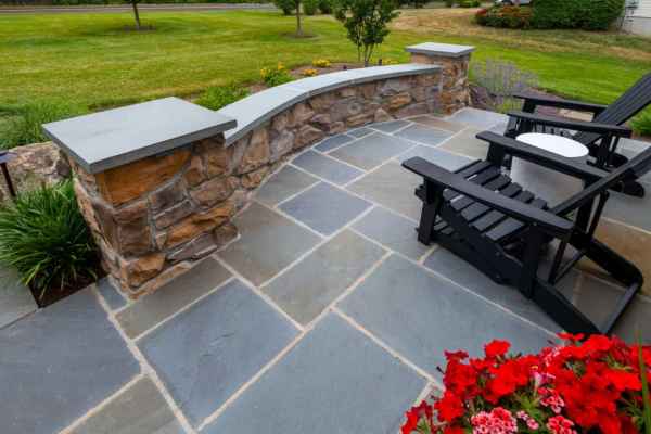 Stone patio with dark wooden chairs and a small round table. A stone wall with a curved design borders the patio, showcasing exceptional hardscaping services in Delaware County, PA. Red flowers in the foreground and a lush green lawn with trees add to the serene outdoor living experience.