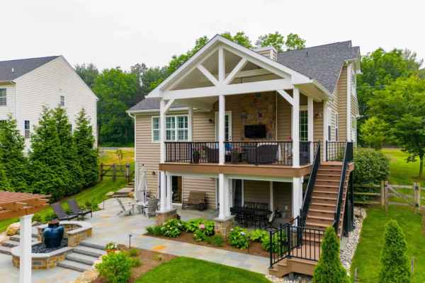 A two-story house in Delaware County, PA offers luxurious outdoor living. The spacious patio boasts a top deck with seating and TV, while below, lounge chairs circle a stone fire pit. Expert landscaping services adorn the yard with greenery and trees, complemented by a wooden staircase connecting the levels.