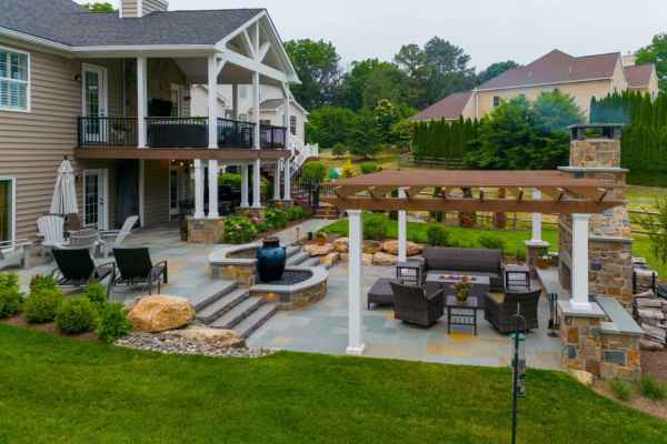 A modern backyard showcasing outdoor living in Delaware County, PA boasts a spacious patio with seating, a pergola, and a fire pit. The area, expertly landscaped with rocks and shrubs, features hardscaping services and includes a water fountain next to the two-story house with balcony furniture.
