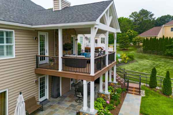A two-story house with a grey roof and covered balcony showcases exquisite outdoor living in Delaware County, PA. The porch features inviting furniture, while the yard boasts lush green grass, elegant bushes, and trees. Stone and siding grace the exterior, complemented by a staircase leading to the balcony.