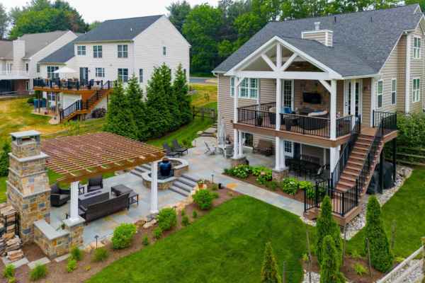 Aerial view of a modern suburban house in Delaware County, PA, with a spacious deck and covered patio. The backyard, crafted by expert landscaping services, features a stone fireplace, pergola, and well-maintained garden. Adjacent houses and lush green trees surround the area.
