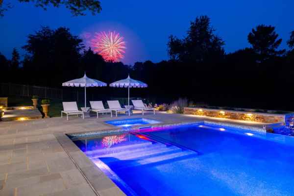 A large, illuminated swimming pool at night with lounge chairs and umbrellas to the side showcases stunning outdoor living. Fireworks burst in the sky behind silhouetted trees in Delaware County, PA, adding vibrant colors to a meticulously designed landscape.