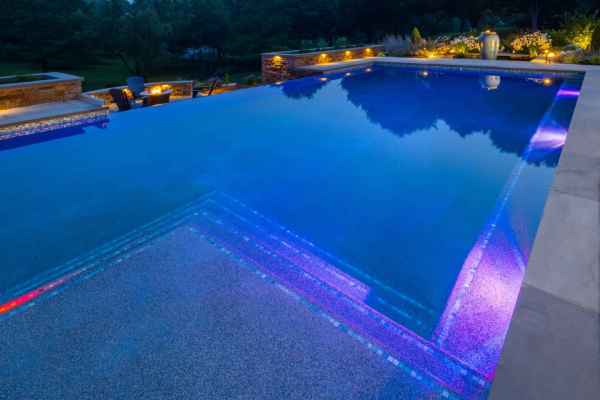 A tranquil swimming pool at night features glowing blue and purple underwater lights. Surrounded by elegant landscape lighting and trees under a starry sky, it offers a serene escape. It's an inviting outdoor living space in Delaware County, PA's finest landscaping tradition.