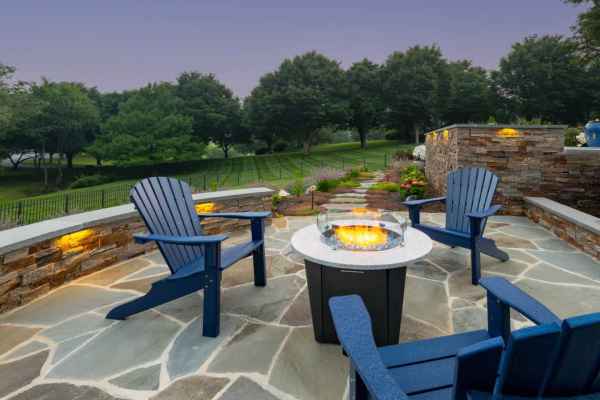 Four blue Adirondack chairs encircle a fire pit on a stone patio during dusk. Warm lights illuminate stone walls, showcasing the exquisite hardscaping services Delaware County PA offers. Beyond, a lush green lawn stretches towards fence and trees as the sky transitions from light purple to dark blue.