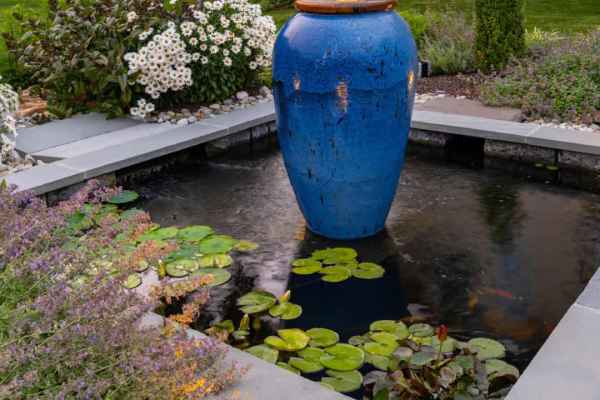 A garden pond with lily pads features a tall, blue ceramic fountain vase at its center. The surrounding landscaping services in Delaware County, PA include flowering bushes, small trees, and a neatly trimmed lawn. A stone path leads to a nearby building with a white door.