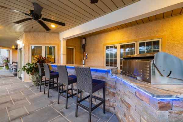 Outdoor living in Delaware County, PA is elevated with a stone bar counter and three woven bar stools. A grill rests under the wooden ceiling, complete with fans. The warm glow of recessed lighting and lush potted plants complete this inviting oasis.