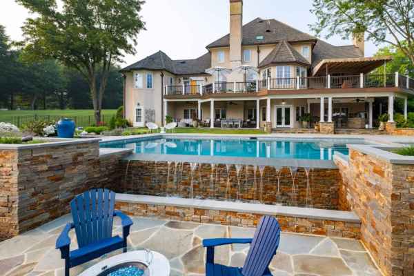 Luxurious house with stone patio features a multi-level pool and waterfall. Blue Adirondack chairs surround a fire pit. The house, enhanced by expert hardscaping services in Delaware County, PA, boasts large windows, balconies, and a spacious outdoor dining area amid lush greenery.