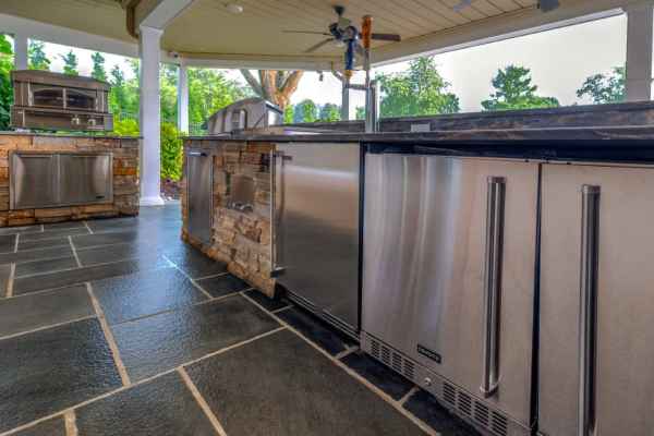 Outdoor living in Delaware County, PA, is epitomized by this kitchen, boasting stainless steel appliances and stone accents. A marble countertop complements an oven, fridge, and tap on a slate tile floor with lush greenery beyond. Ideal for those seeking exquisite hardscaping services.