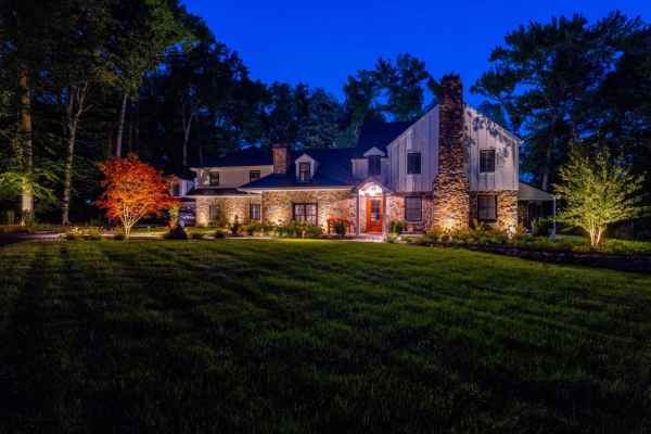 A charming house with a mix of stone and siding exterior, warmly lit at night. Surrounded by trees and expert hardscaping services in Delaware County, PA, it boasts a well-manicured lawn and an illuminated red-leaved tree near the entrance, creating a cozy, inviting atmosphere.