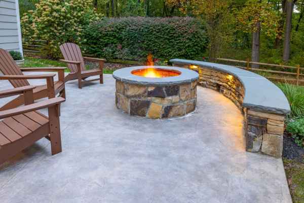 A cozy outdoor patio with a stone fire pit in the center is surrounded by wooden chairs and a curved stone bench with built-in lighting. The area, crafted through expert hardscaping services in Delaware County PA, is bordered by lush greenery and trees.