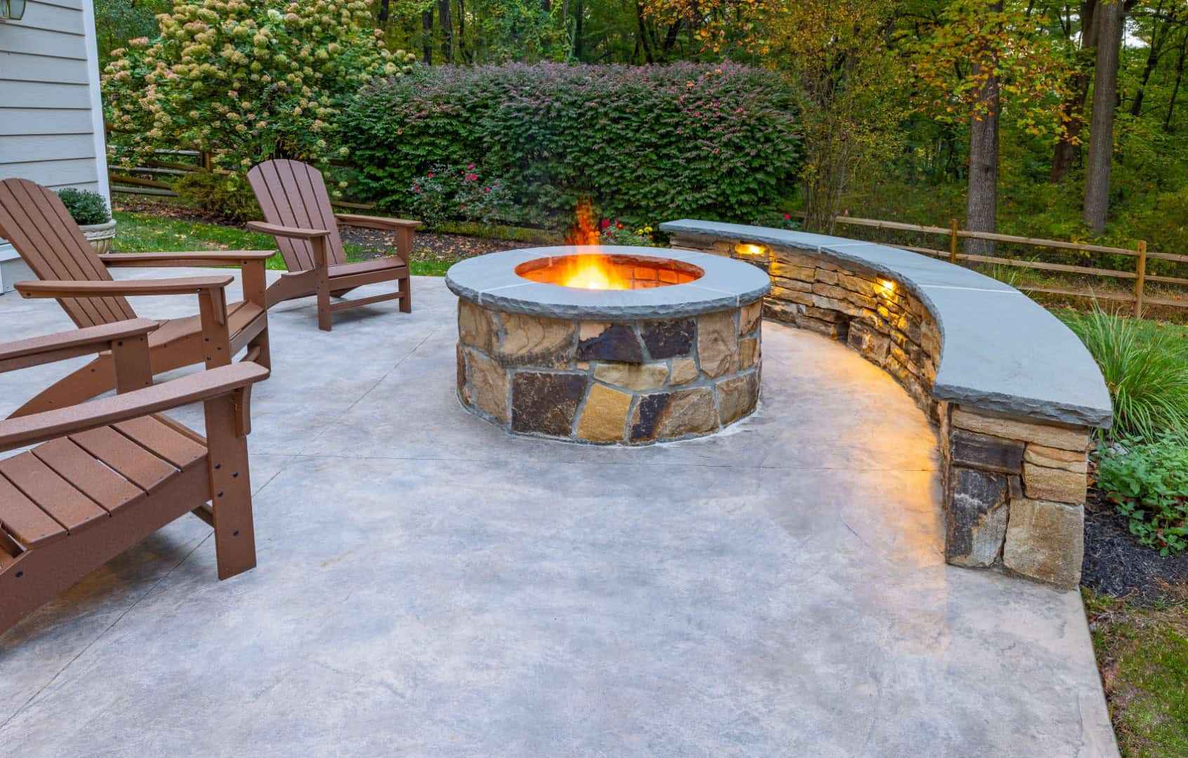 A cozy outdoor patio in Delaware County, PA, features a stone fire pit with orange flames, surrounded by wooden Adirondack chairs. Soft lighting highlights a curved stone bench at the edge, framed by lush greenery and trees—perfect for those seeking exceptional hardscaping services.