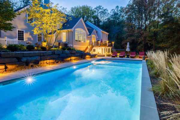 A well-lit, modern house with large windows is set against a backdrop of trees during dusk. A rectangular swimming pool with glowing lights reflects the house, showcasing exquisite outdoor living in Delaware County, PA. Red lounge chairs and a large umbrella are by the poolside.