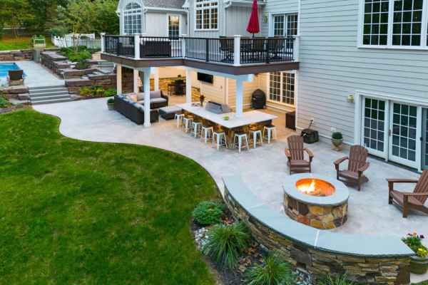 A spacious backyard in Delaware County, PA, features a white house with a large patio area. With exquisite outdoor living elements like cushioned seating and a fire pit surrounded by Adirondack chairs, lush landscaping enhances the green grass while a pool glistens in the background.