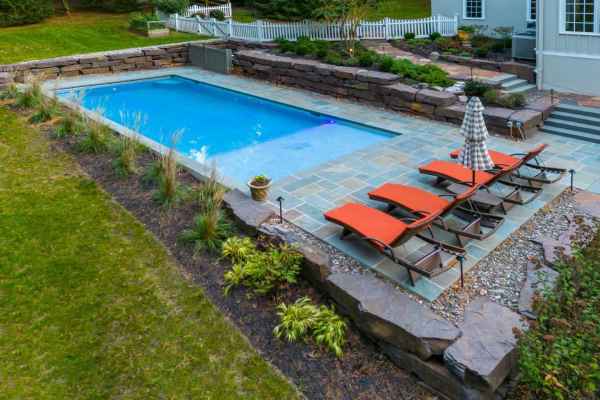 A backyard scene with a rectangular swimming pool surrounded by a stone patio showcases exceptional outdoor living Delaware County PA. Four orange lounge chairs and a striped umbrella rest beside the pool, bordered by lush landscaping and a classic stone wall.