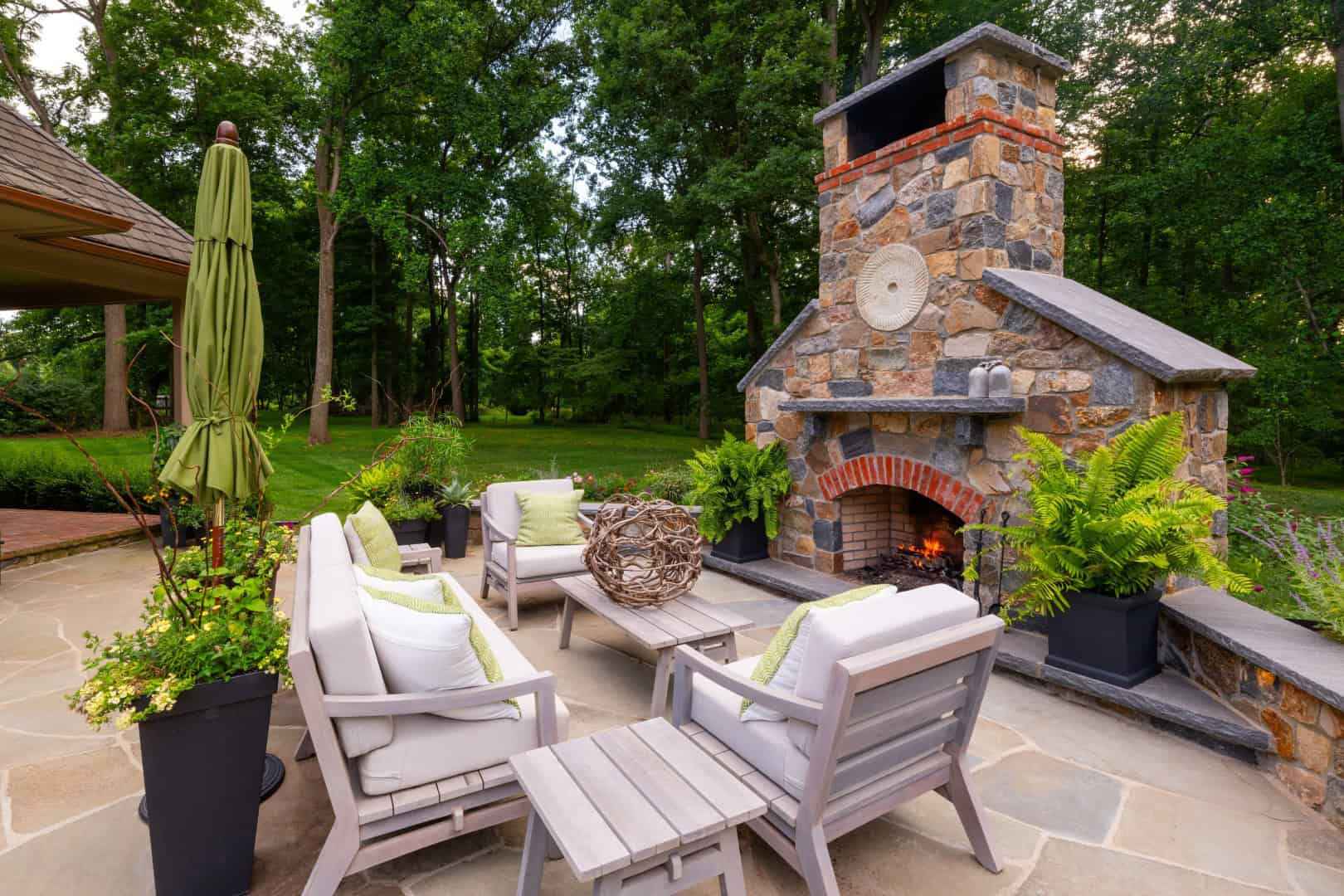 Outdoor patio with wooden furniture arranged around a lit stone fireplace. Green cushions accent the chairs, and potted plants add greenery. An umbrella stands nearby, with lush trees in the background. This cozy outdoor living setting highlights premier hardscaping services in Delaware County, PA.