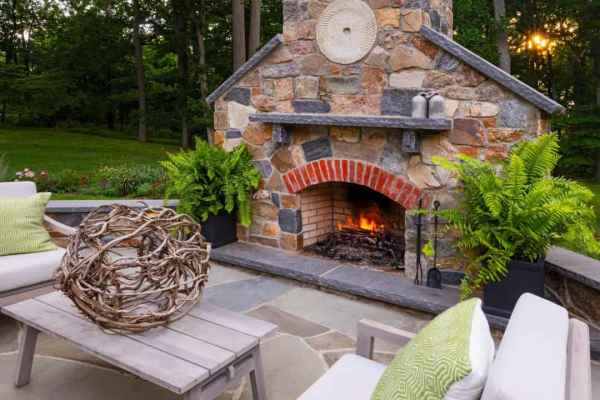 Experience elevated outdoor living in Delaware County, PA, with a stone fireplace surrounded by lush greenery. Comfy chairs and green pillows create a cozy ambiance. A decorative wicker ball graces the wooden coffee table, making this an ideal retreat enhanced by expert landscaping services.