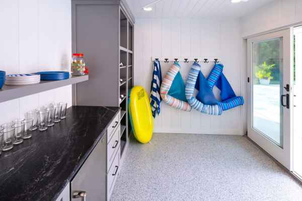 A bright poolside room with towels hung on hooks, a yellow float leaning against the wall, and various plates and glasses on a countertop. Sunlight streams in through a sliding glass door to reveal exquisite outdoor living crafted by top landscaping services in Delaware County, PA.