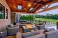 A cozy outdoor patio features wicker furniture with gray cushions, centered around a modern fire pit. A wooden pergola roof with skylights covers the area. This serene outdoor living setup in Delaware County, PA, is complemented by a manicured lawn and trees, with lounge chairs on a nearby deck.
