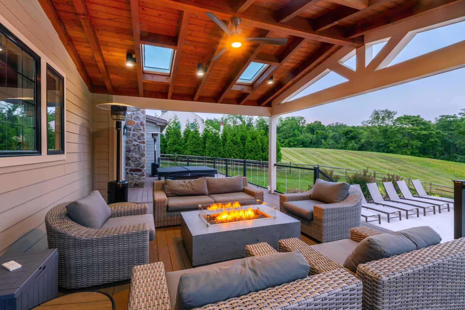 A cozy outdoor patio, perfect for enjoying Delaware County's finest outdoor living, features wicker furniture with gray cushions around a rectangular fire pit. A wooden ceiling with skylights covers the space, while a grassy lawn and neatly lined lounge chairs complete this serene setting.