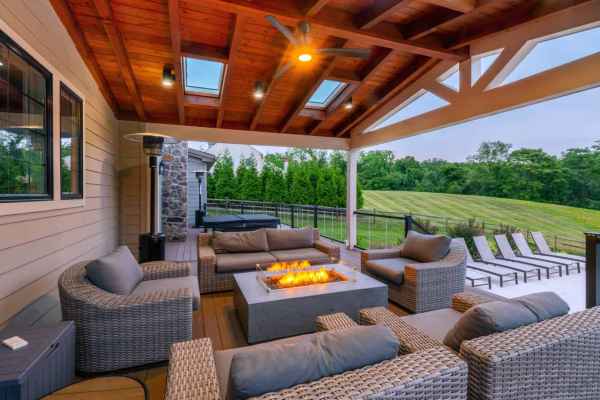 A cozy outdoor patio, perfect for enjoying Delaware County's finest outdoor living, features wicker furniture with gray cushions around a rectangular fire pit. A wooden ceiling with skylights covers the space, while a grassy lawn and neatly lined lounge chairs complete this serene setting.