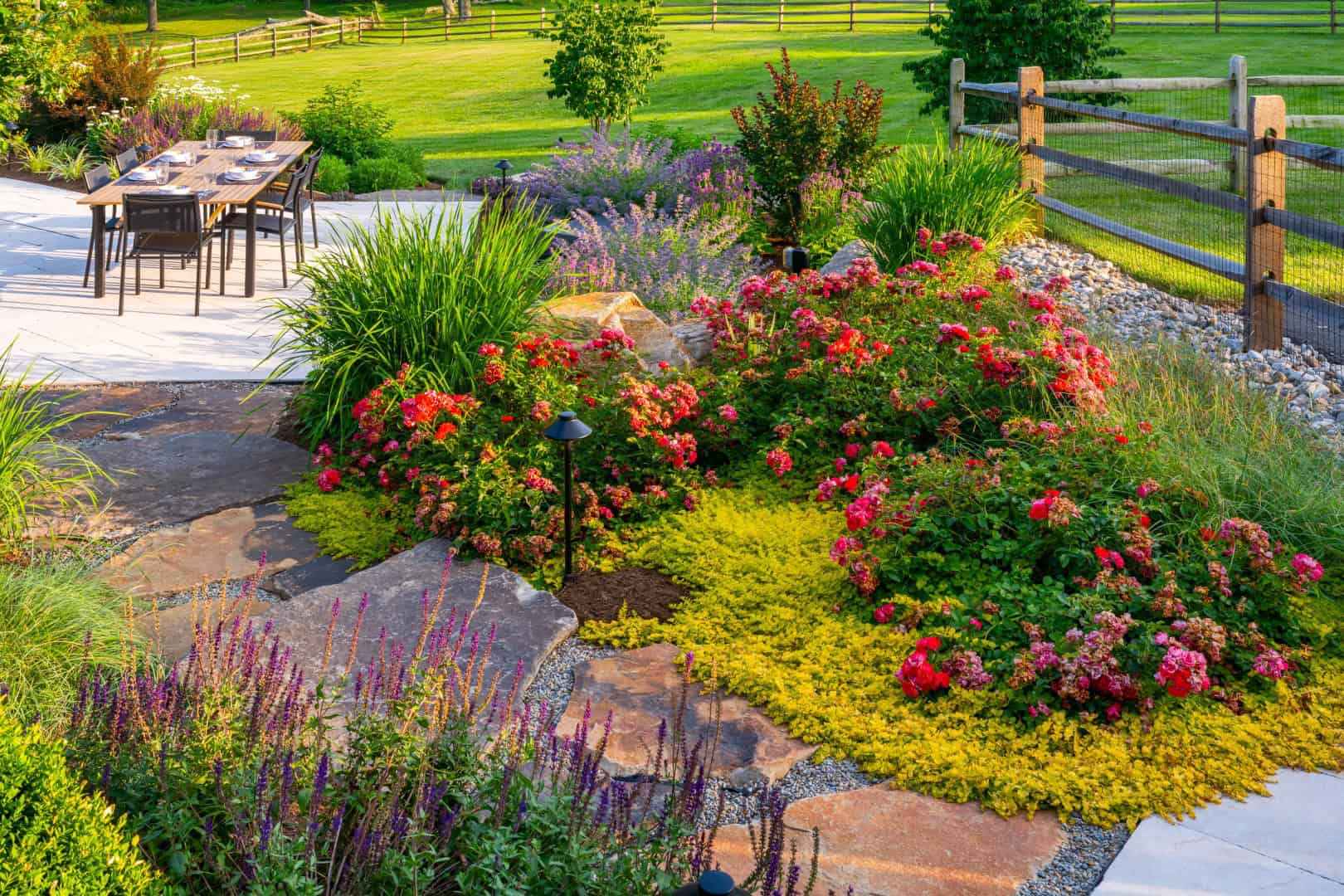 A landscaped garden with vibrant red and pink flowers is surrounded by lush greenery in Delaware County, PA. A wooden fence encloses the area, where a patio with a table and chairs offers the perfect outdoor living experience, set against a sunny, green lawn backdrop.