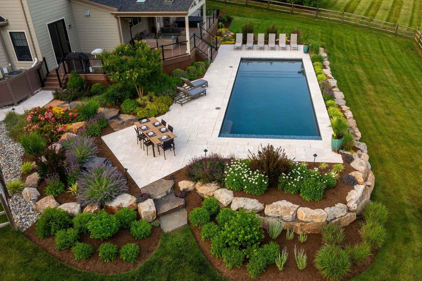 Aerial view of a backyard with a rectangular pool, surrounded by an expertly crafted stone patio, showcases premium hardscaping services in Delaware County, PA. Lounge chairs and a dining set complement the lush gardens adorned with colorful plants and rocks encircling the area.