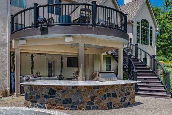 Outdoor living in Delaware County, PA, shines with a round, elevated deck featuring sleek black railings. Below, a stone bar and grill invite gatherings. Curved stairs ascend to the deck, while large windows and light-colored walls of the house complete this stunning hardscape.