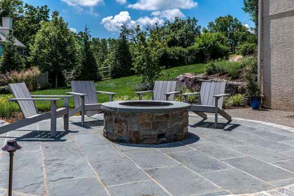 Outdoor living in Delaware County, PA, features a serene patio with gray Adirondack chairs encircling a stone fire pit on slate tiles. The lush greenery and blue sky create the perfect backdrop for relaxation.