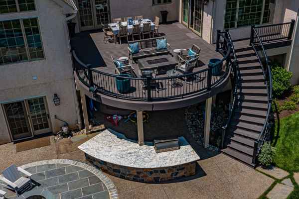 Aerial view of a modern two-story patio with elegant outdoor furniture. The upper deck boasts a dining area and lounge chairs surrounding a fire pit. A curved staircase leads to a lower stone patio with built-in barbecue and seating—perfect for embracing outdoor living in Delaware County PA.