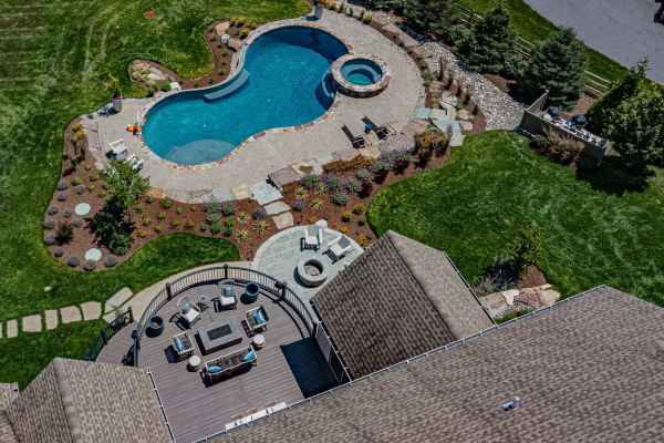 Aerial view of a landscaped backyard in Delaware County, PA, showcasing a curvy swimming pool with a hot tub. Adjacent is a circular stone fire pit surrounded by furniture on a wooden deck. This outdoor living space features well-manicured lawns and pathways, perfect for relaxation.