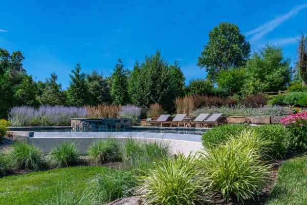 Beautiful backyard pool surrounded by lush greenery, colorful flowers, and tall bushes. Four wooden lounge chairs are arranged on a stone deck beside the pool. A waterfall feature adds to the tranquil scene under a clear blue sky, showcasing expert landscaping services in Delaware County, PA.