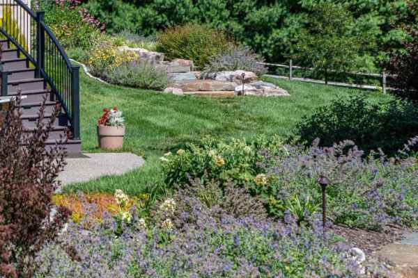 A landscaped garden with a stone path leading uphill, surrounded by blooming flowers and lush greenery. A pot of red flowers sits near the steps of a house on the left. With expert landscaping services in Delaware County, PA, enjoy trees and a wooden fence gracing the background.