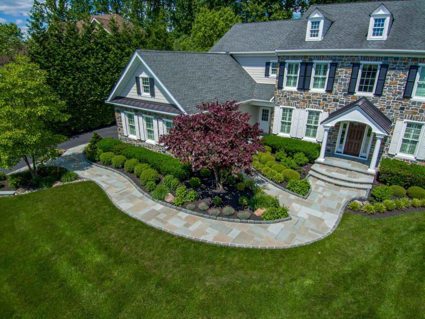 Aerial view of a large house with stone and white siding. The yard showcases expert landscaping services in Delaware County, PA, featuring a well-maintained garden with a central red-leafed tree and green shrubs. A stone pathway enhances the outdoor living space, leading to the entrance amidst lush grass.